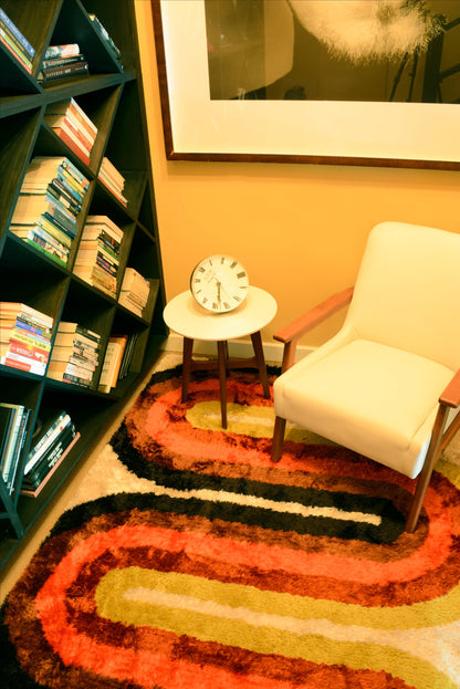 Cozy retro reading nook: dark wood bookshelf, cream armchair, round side table with clock, and a multicolored shag runner rug.