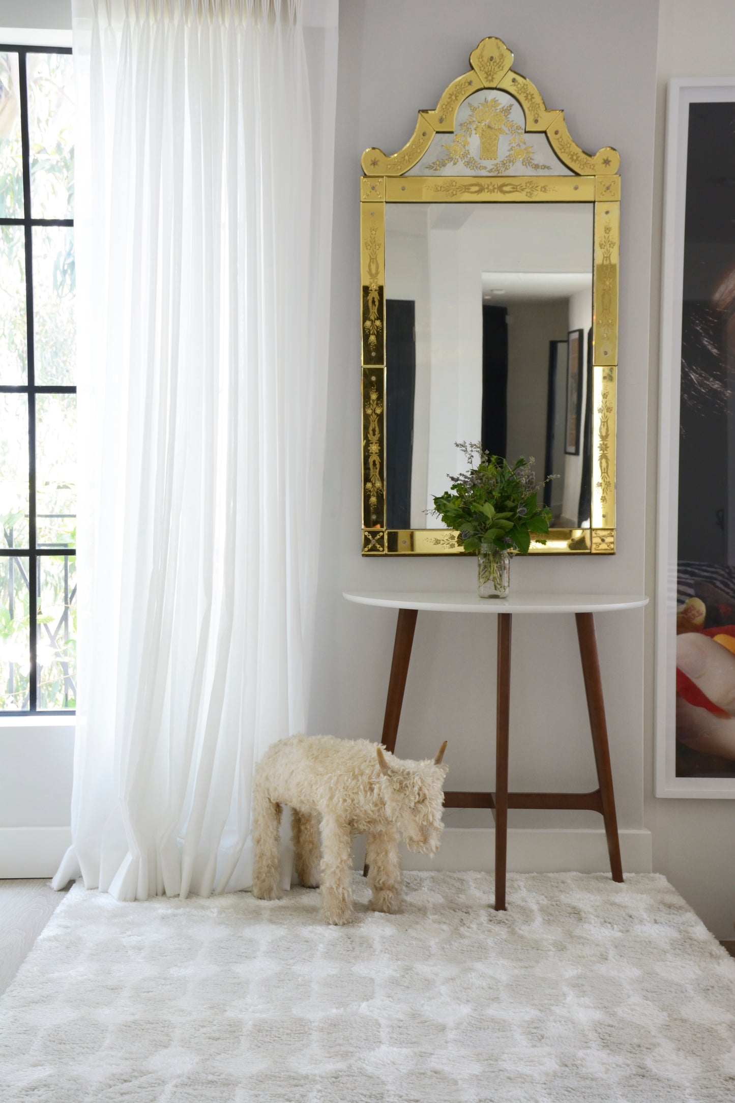 Retro gold ornate rug on a living room floor, with a white round console table on wooden legs, a plant in a vase, a fluffy dog statue, and sheer curtains.