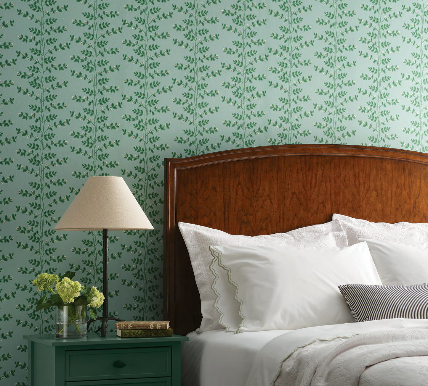 Bedroom with soft teal climbing-vine wallpaper, dark wood headboard, white bedding, nightstand, lampshade, and a vase of yellow flowers.