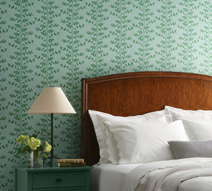 Bedroom with soft teal climbing-vine wallpaper, dark wood headboard, white bedding, nightstand, lampshade, and a vase of yellow flowers.