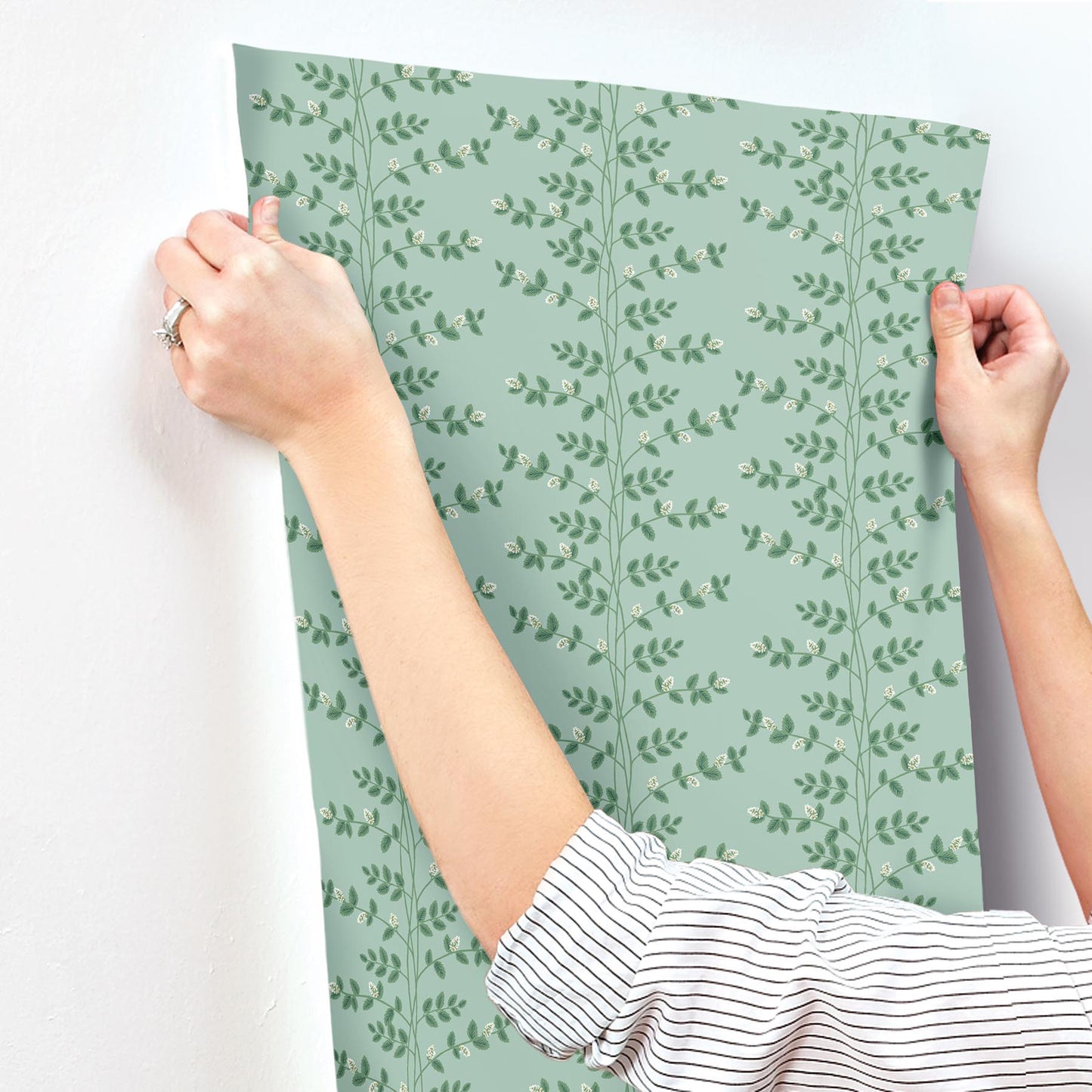 Soft teal wallpaper with a vertical leafy vine pattern; hands smooth the sheet onto a wall, sample swatch held.