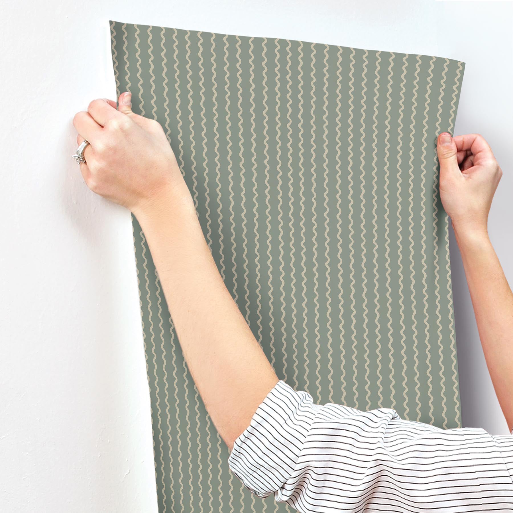 Grey-green wallpaper panel with moss-green Rickrack stripe pattern, held by two hands in a striped shirt.