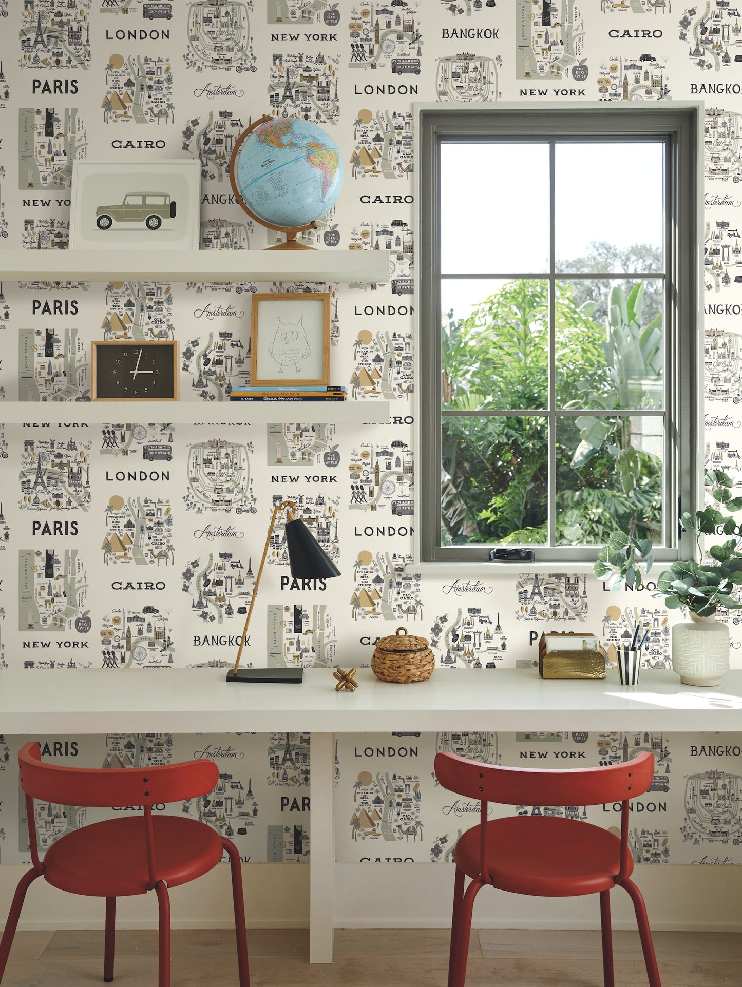 Travel-inspired home office with white desk and two red chairs, city maps grey and gold wallpaper, globe, frames, lamp, window and plants.