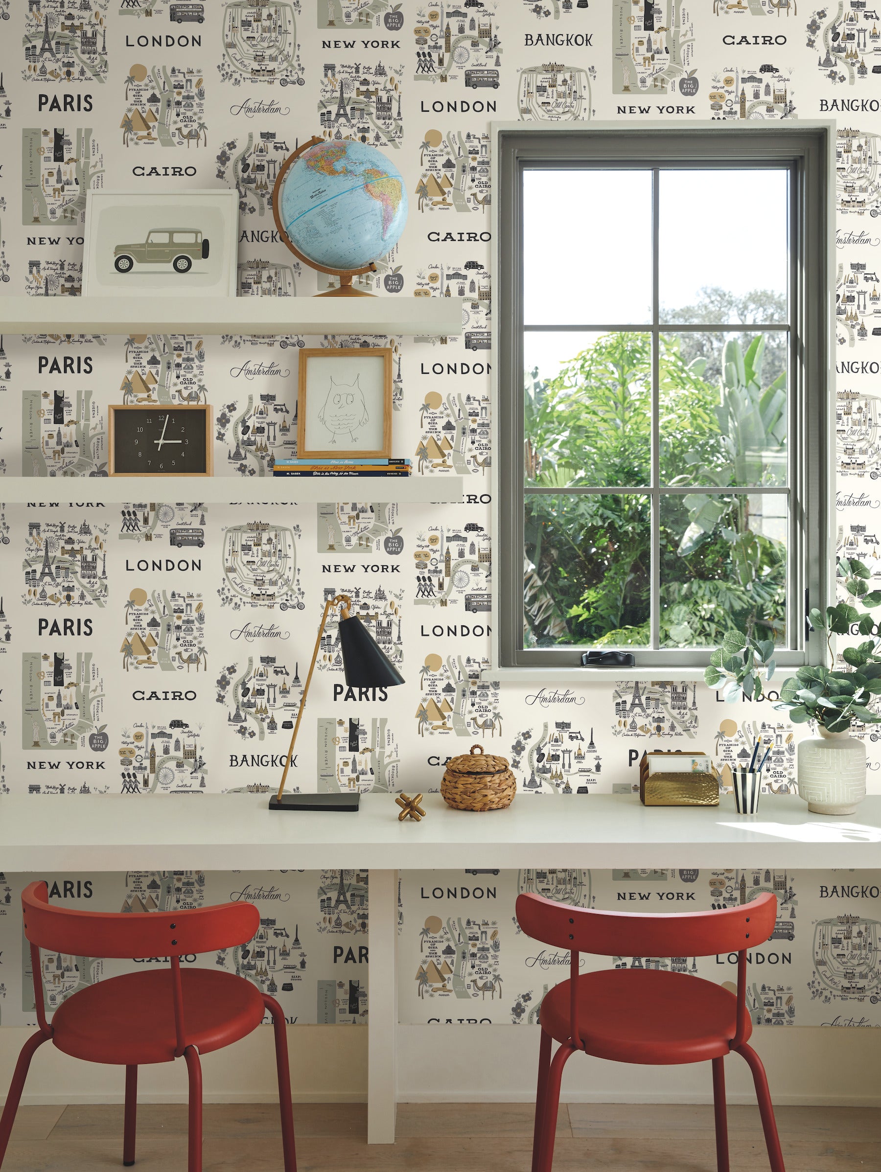 Travel-inspired home office with white desk and two red chairs, city maps grey and gold wallpaper, globe, frames, lamp, window and plants.