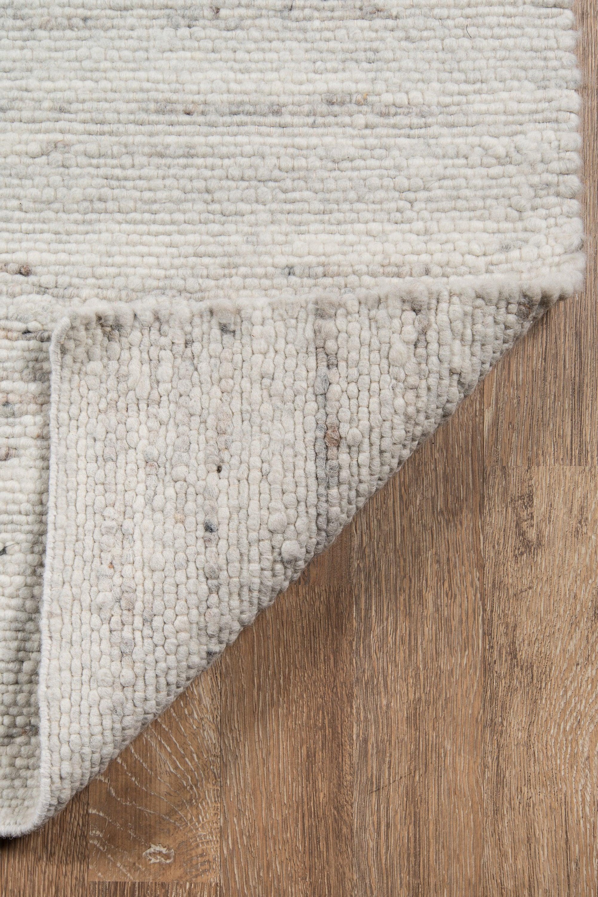 Ivory textured loop rug with thick pile, folded corner to reveal underside, on warm wooden floor.