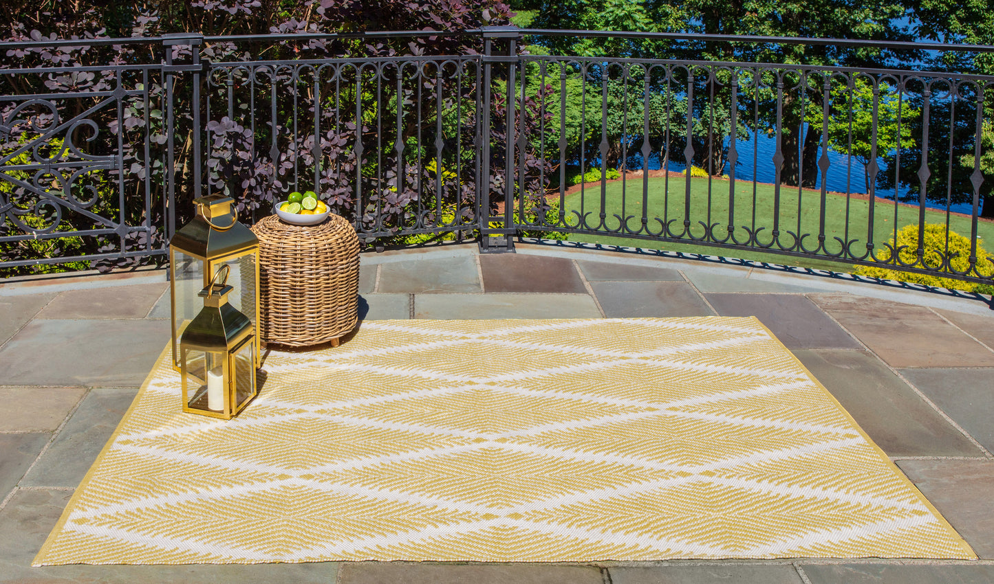 Yellow and white geometric outdoor rug on stone patio with brass lanterns, wicker side table with limes, and black railing.