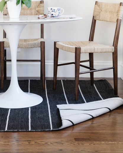 Mid-century white tulip table with round top and two dark wood chairs with woven seats on striped rug; teacup and glasses.