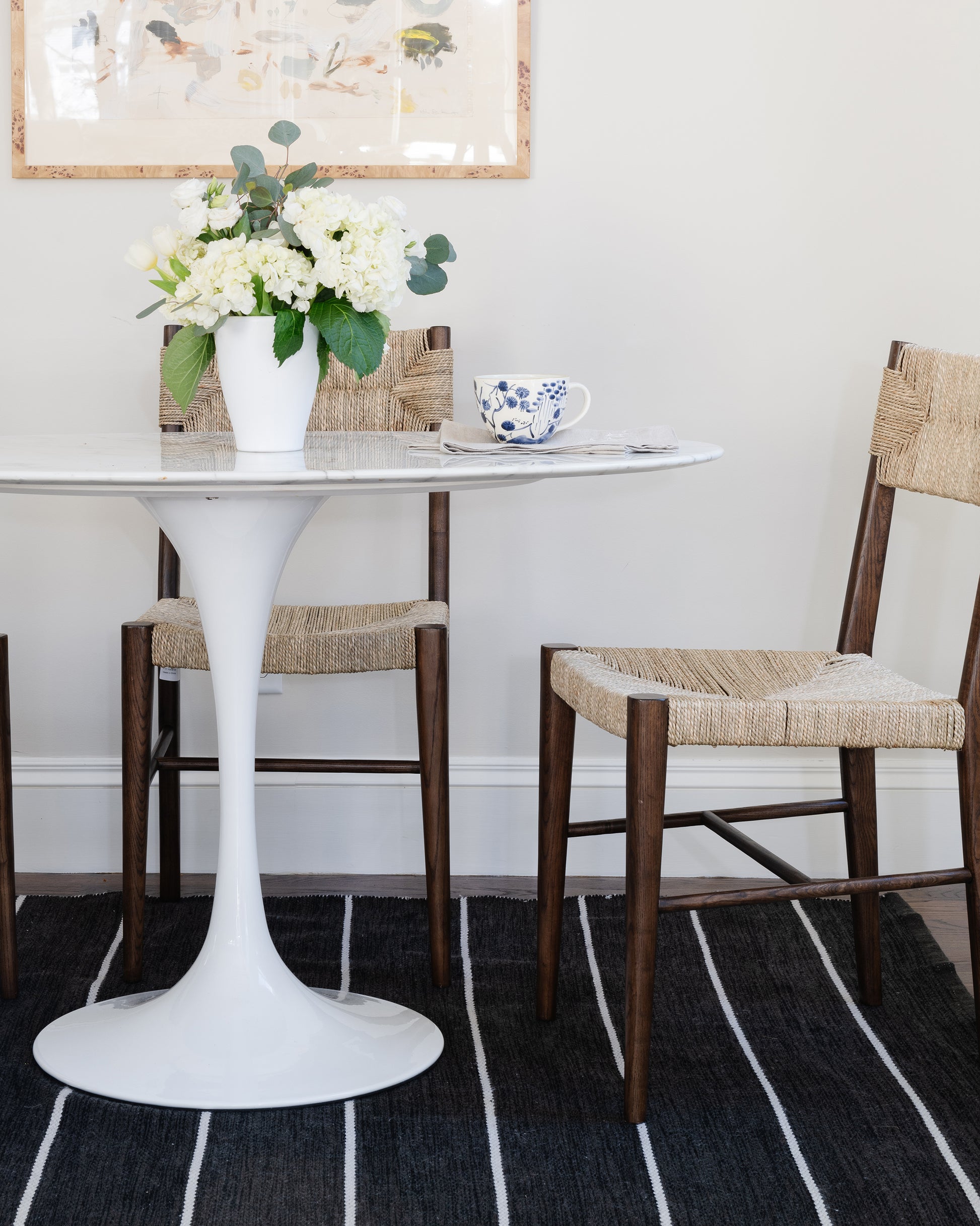 White round pedestal table with marble top, wooden wicker chairs, vase of white flowers, blue floral teacup, striped rug.