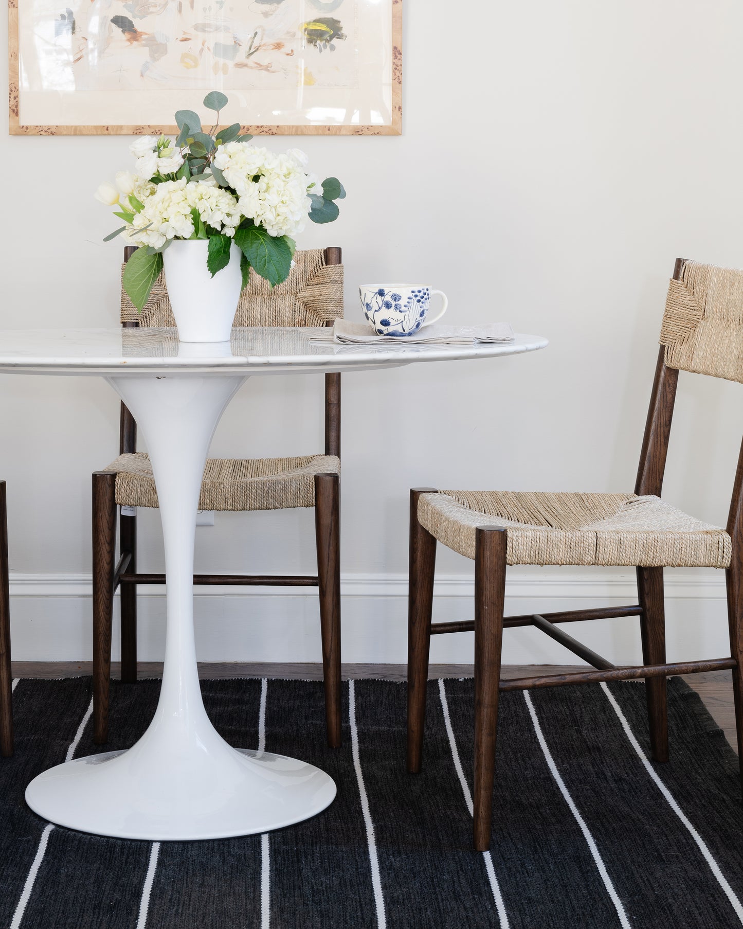 White round marble-top pedestal table & two woven-seat wooden chairs, vase of white flowers, blue teacup, framed art.