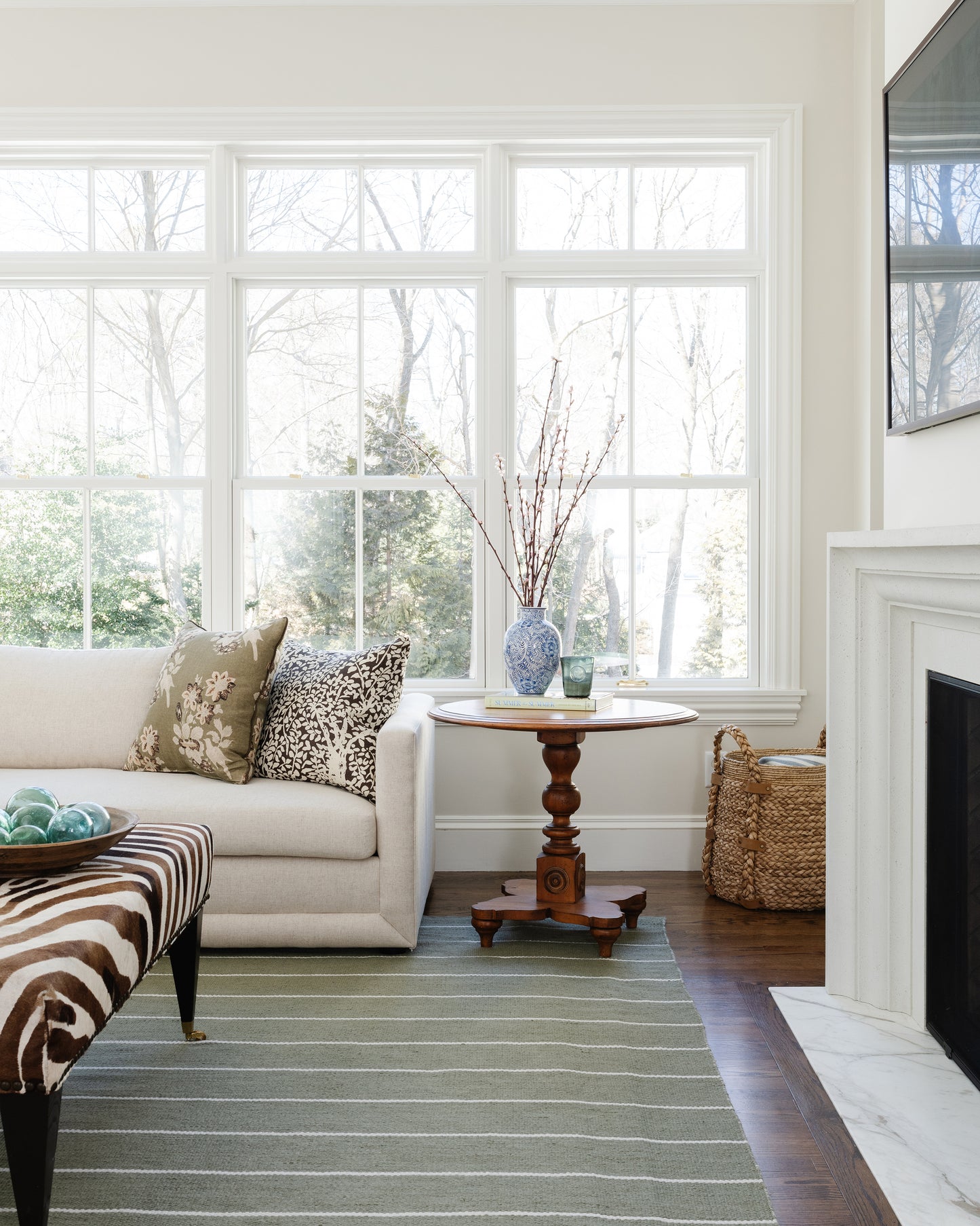Bright living room with cream sofa, zebra ottoman, round wood table, blue vase with twigs, woven basket, fireplace, windows.