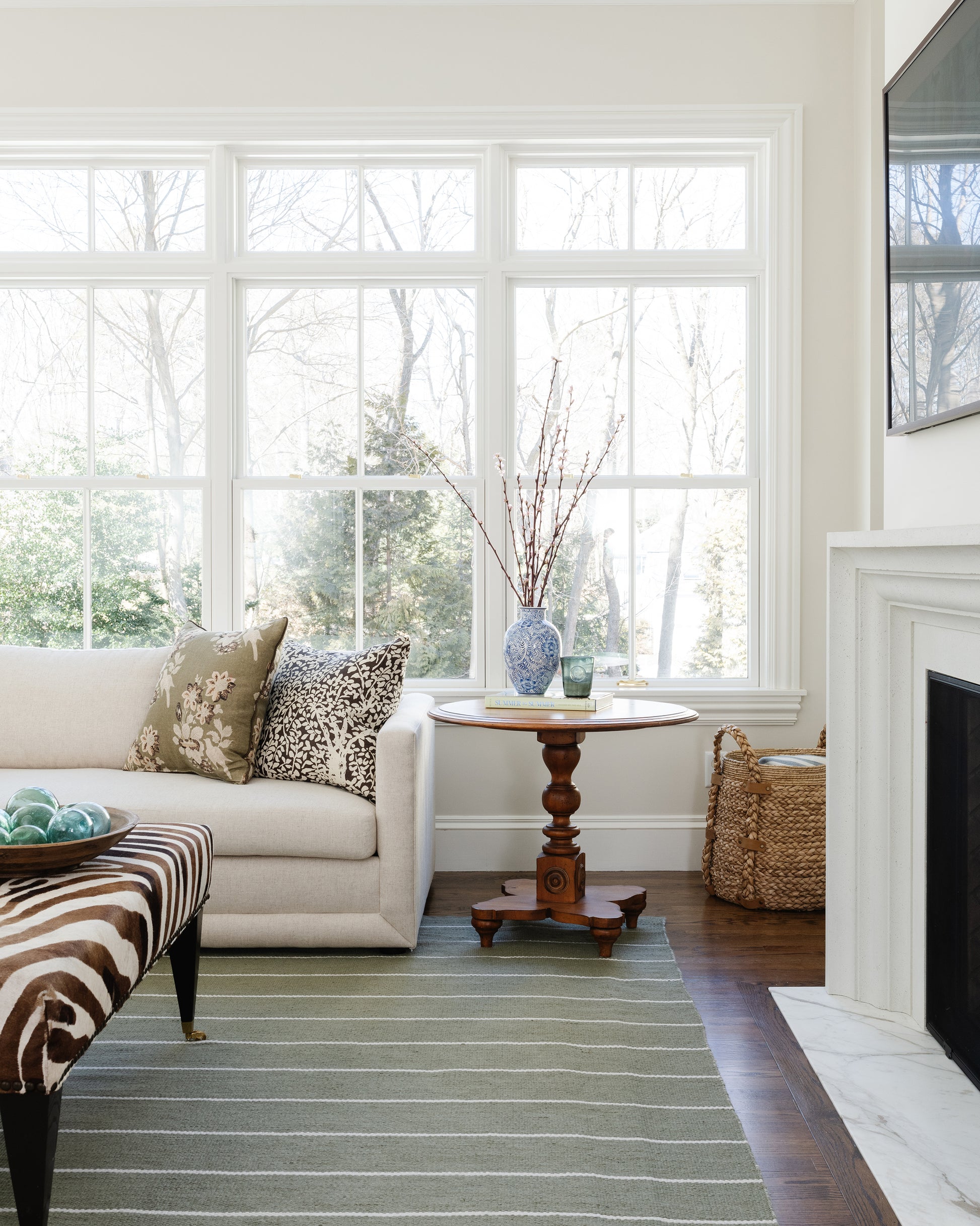 Bright living room with cream sofa, zebra ottoman, round wood table, blue vase with twigs, woven basket, fireplace, windows.
