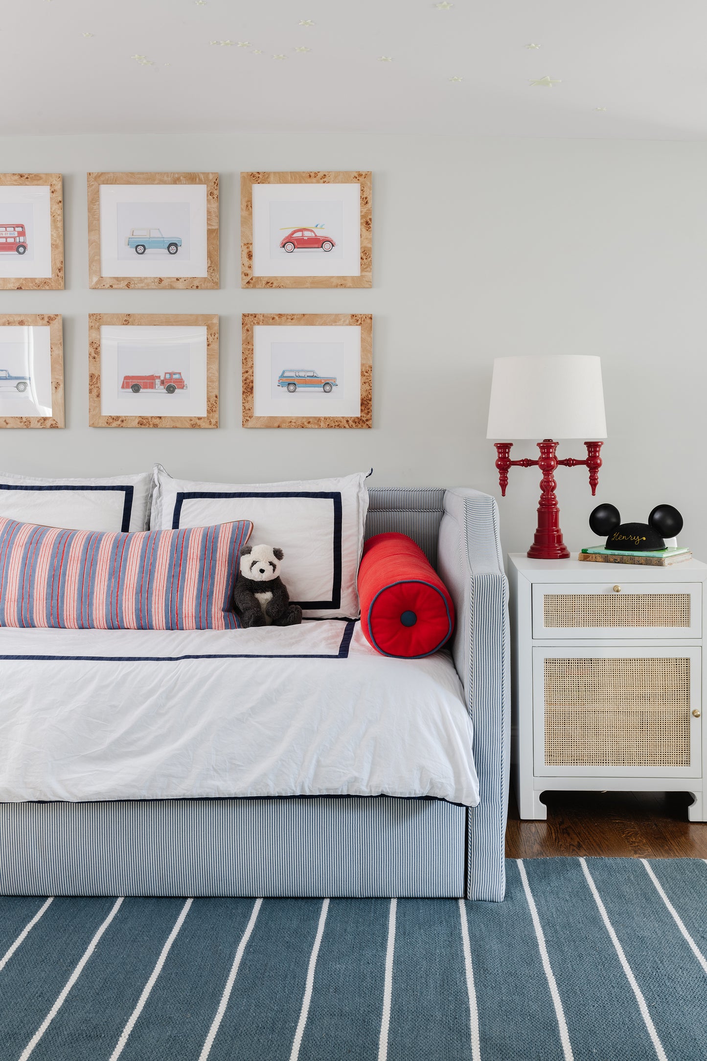 Coastal twin bed with white duvet and navy piping, striped lumbar pillow, red bolster; cork-framed car art on wall.