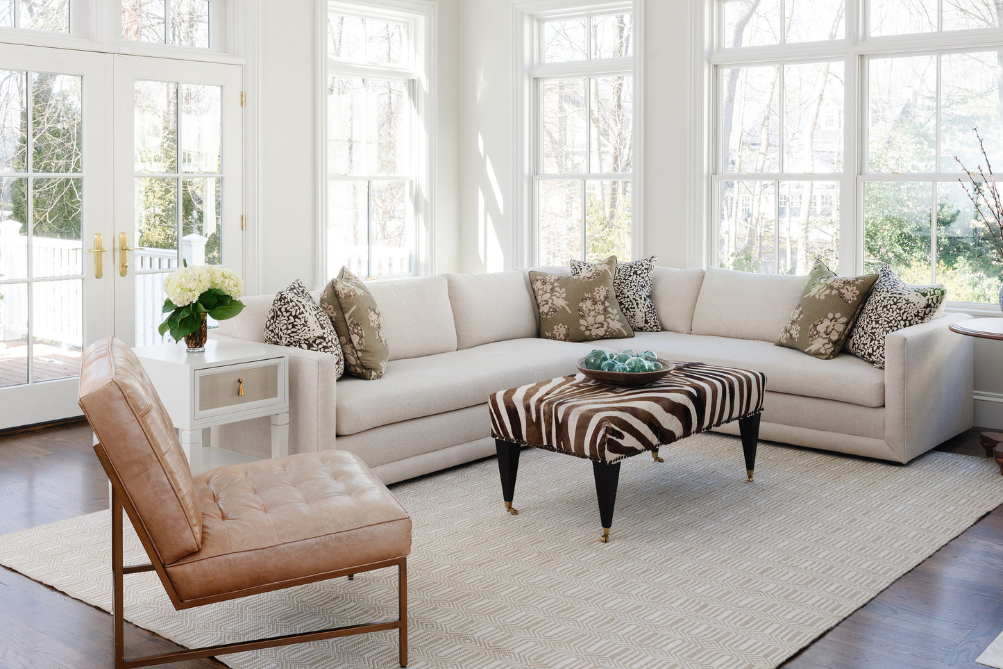 Bright living room scene with a beige sectional, zebra ottoman, animal-print pillows, tan leather chair, white side table, and a rug on the floor.