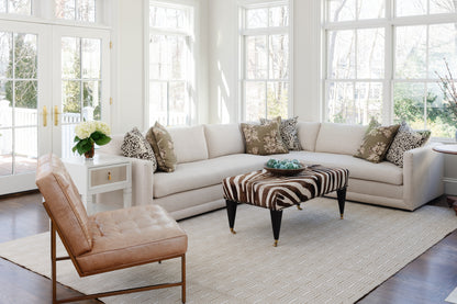 Bright living room scene with a beige sectional, zebra ottoman, animal-print pillows, tan leather chair, white side table, and a rug on the floor.