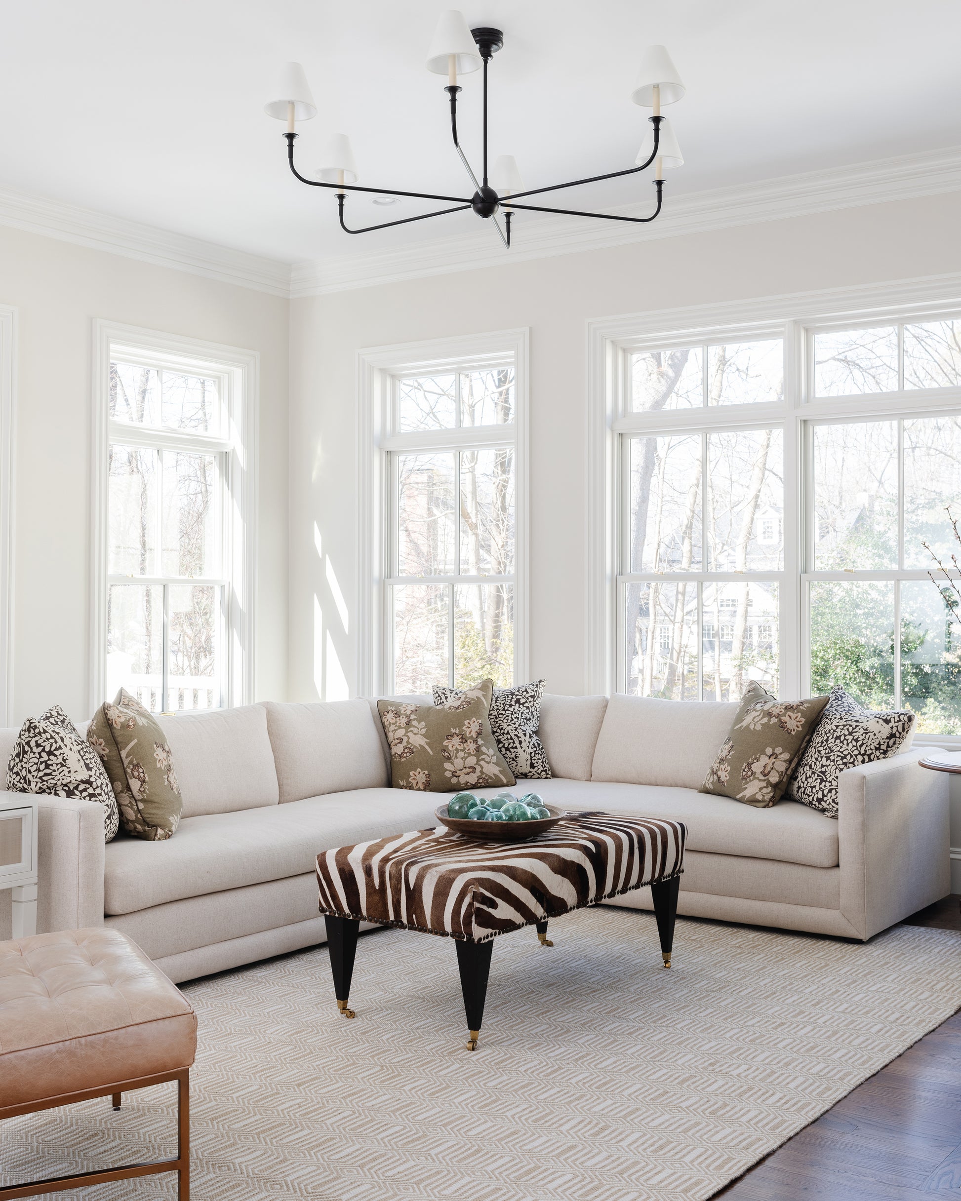 Cream L-shaped sofa with zebra-print ottoman, beige rug & black metal chandelier in a bright sunlit living room.