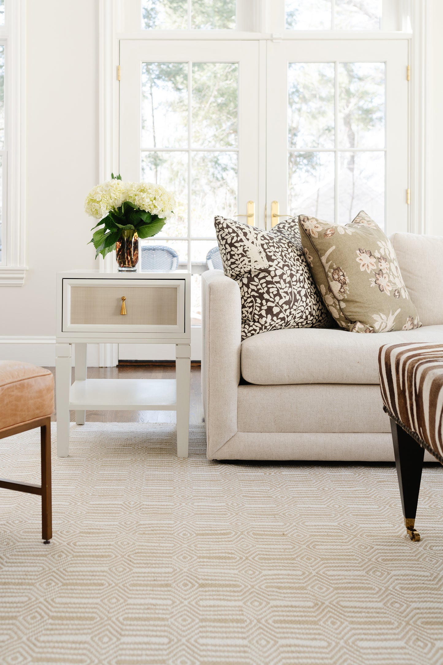 Neutral living room with beige sofa, geometric rug, patterned pillows, gray end table with gold hardware, white flowers.