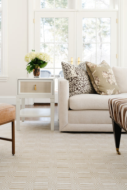Neutral living room with beige sofa, geometric rug, patterned pillows, gray end table with gold hardware, white flowers.