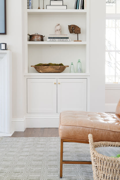 Green rug 2' x 3' on the floor in a living area with white built-in shelving, decor (wooden bowl, moss centerpiece, glass bottles) and a tan tufted leather ottoman.