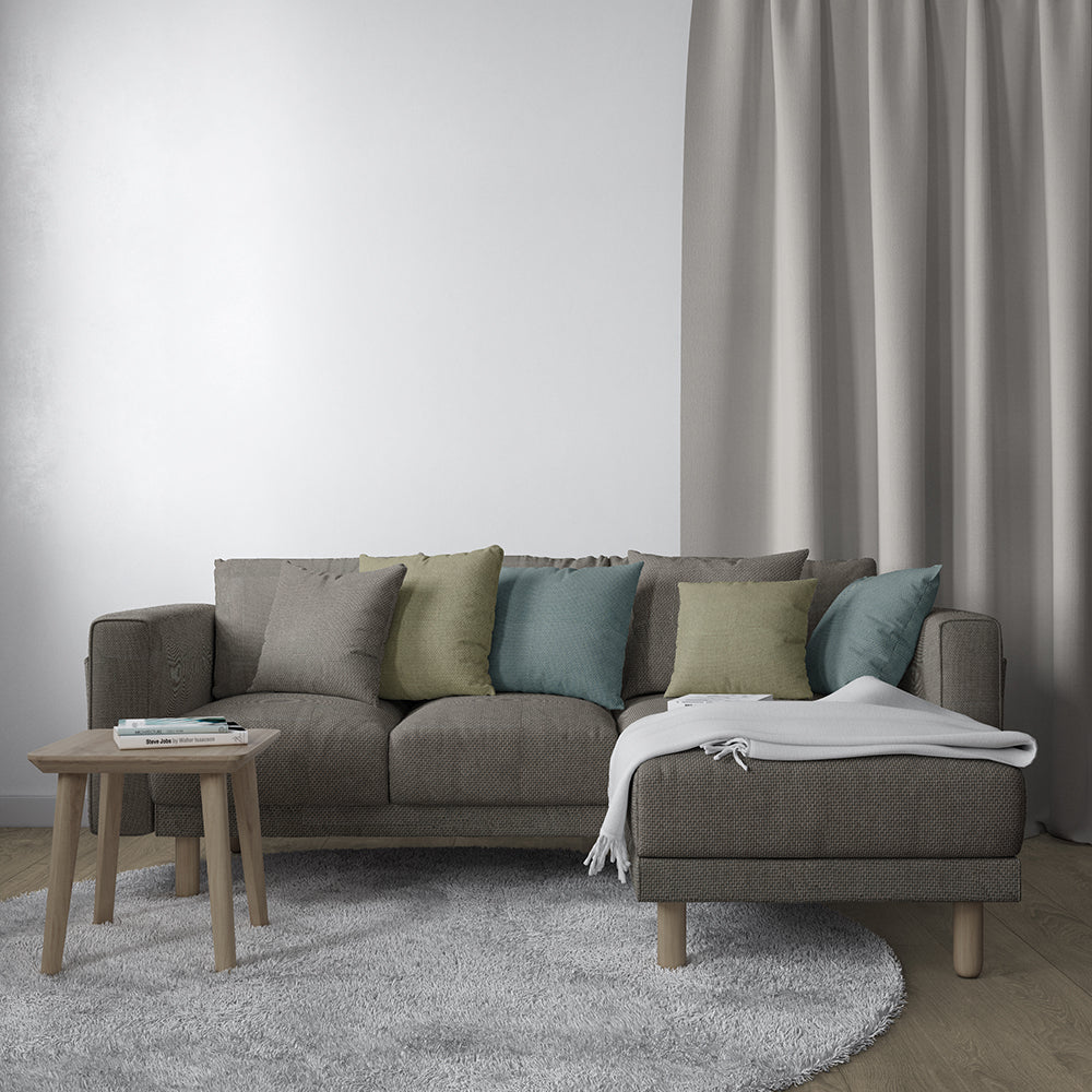 Taupe fabric L-shaped sofa with chaise, sage, teal and beige cushions, light wood legs, round coffee table on gray rug.