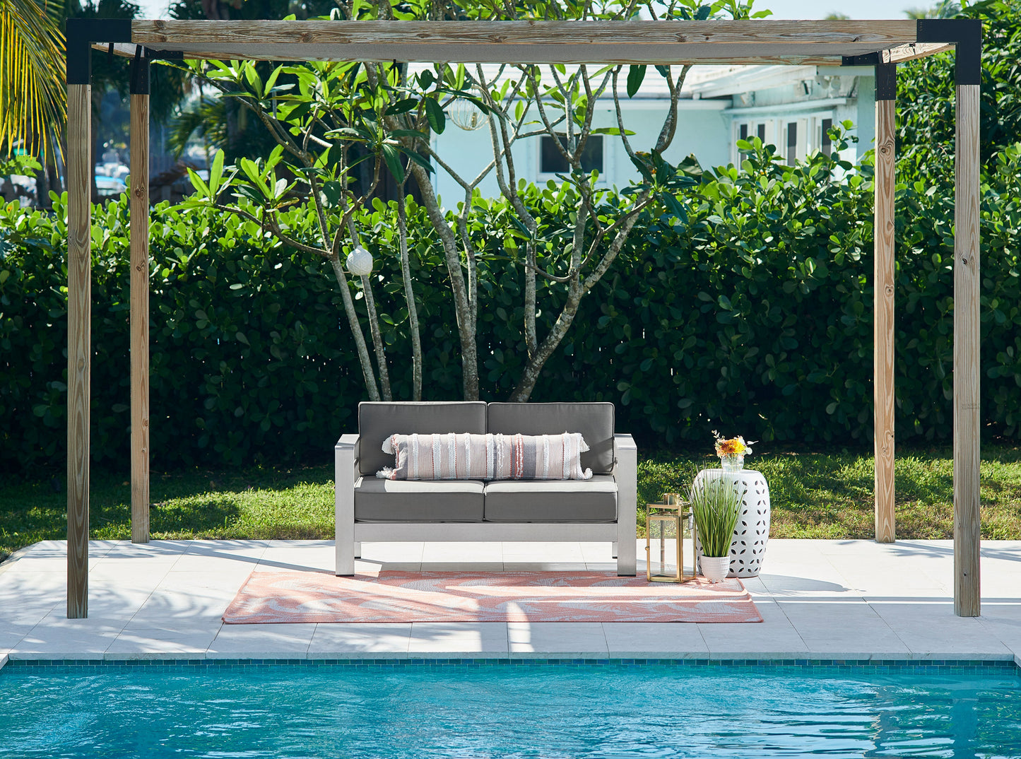Pink outdoor rug on a poolside patio under a wooden pergola with greenery and a vase.