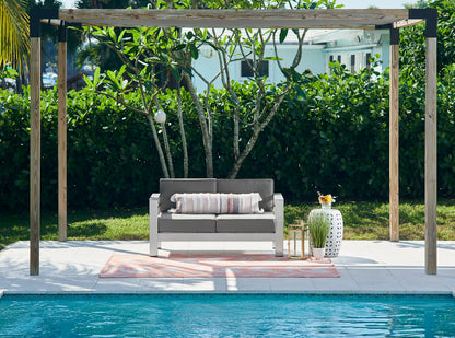 Peach outdoor area rug (7' x 9') under a wooden pergola beside a pool; grey outdoor loveseat with a striped cushion sits on the rug, with a white vase and lush hedges nearby.