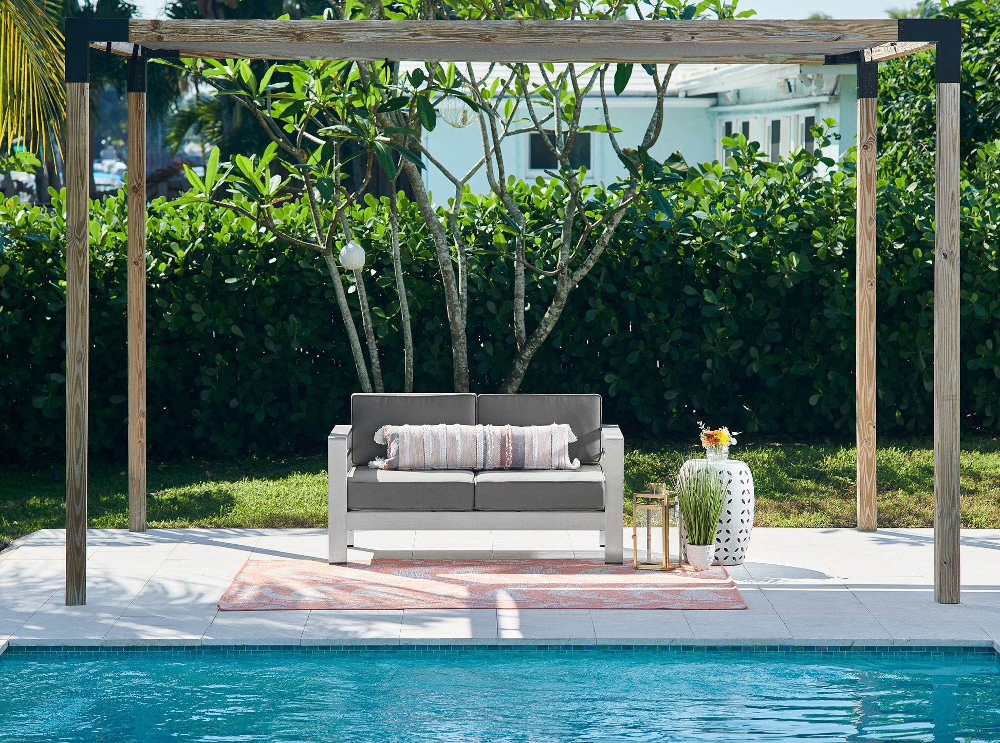 Outdoor poolside seating under a wooden pergola: gray cushioned loveseat, striped pillows, peach rug, white lattice vase.