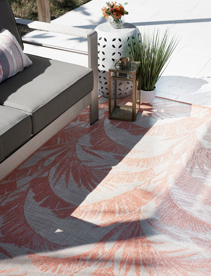 Outdoor patio with gray cushioned metal sofa, coral palm-leaf rug, white side table, gold lantern and potted grasses.
