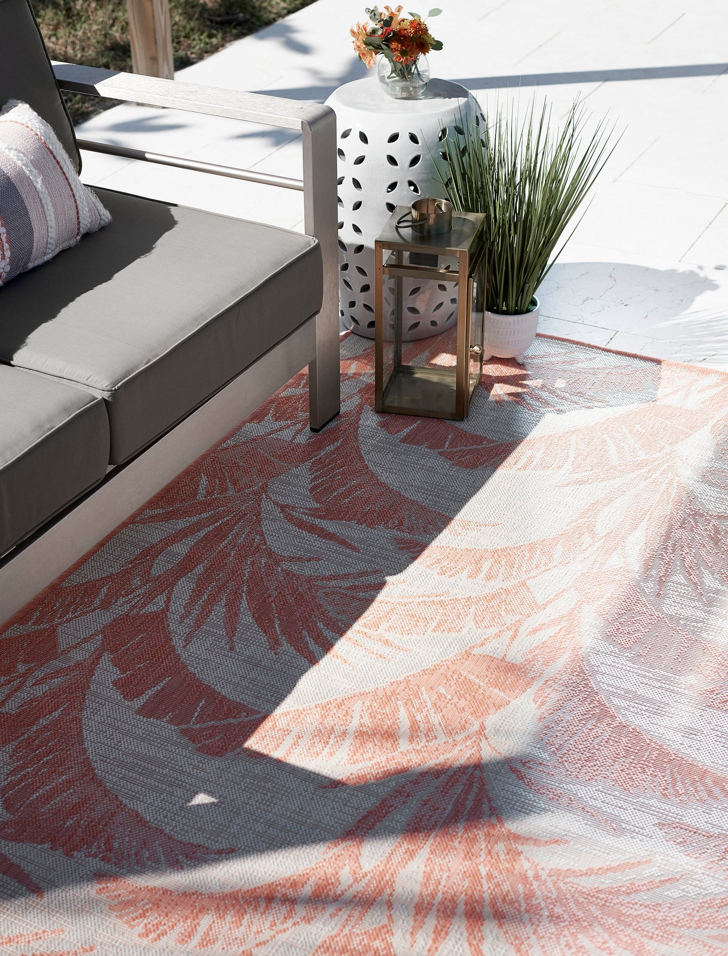 Outdoor patio scene with gray cushioned sofa, peach palm-leaf rug, white lattice lantern and tall grass plant.