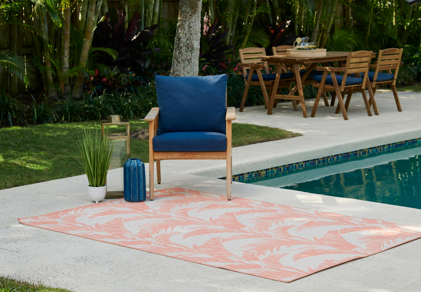Outdoor pool patio: wooden armchair with blue cushions, peach palm-pattern rug, blue vase and lantern beside a dining set.