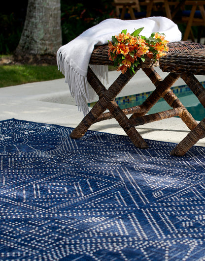 Outdoor blue geometric rug with white dotted pattern, draped white fringed throw over rustic wicker bench, orange flowers.