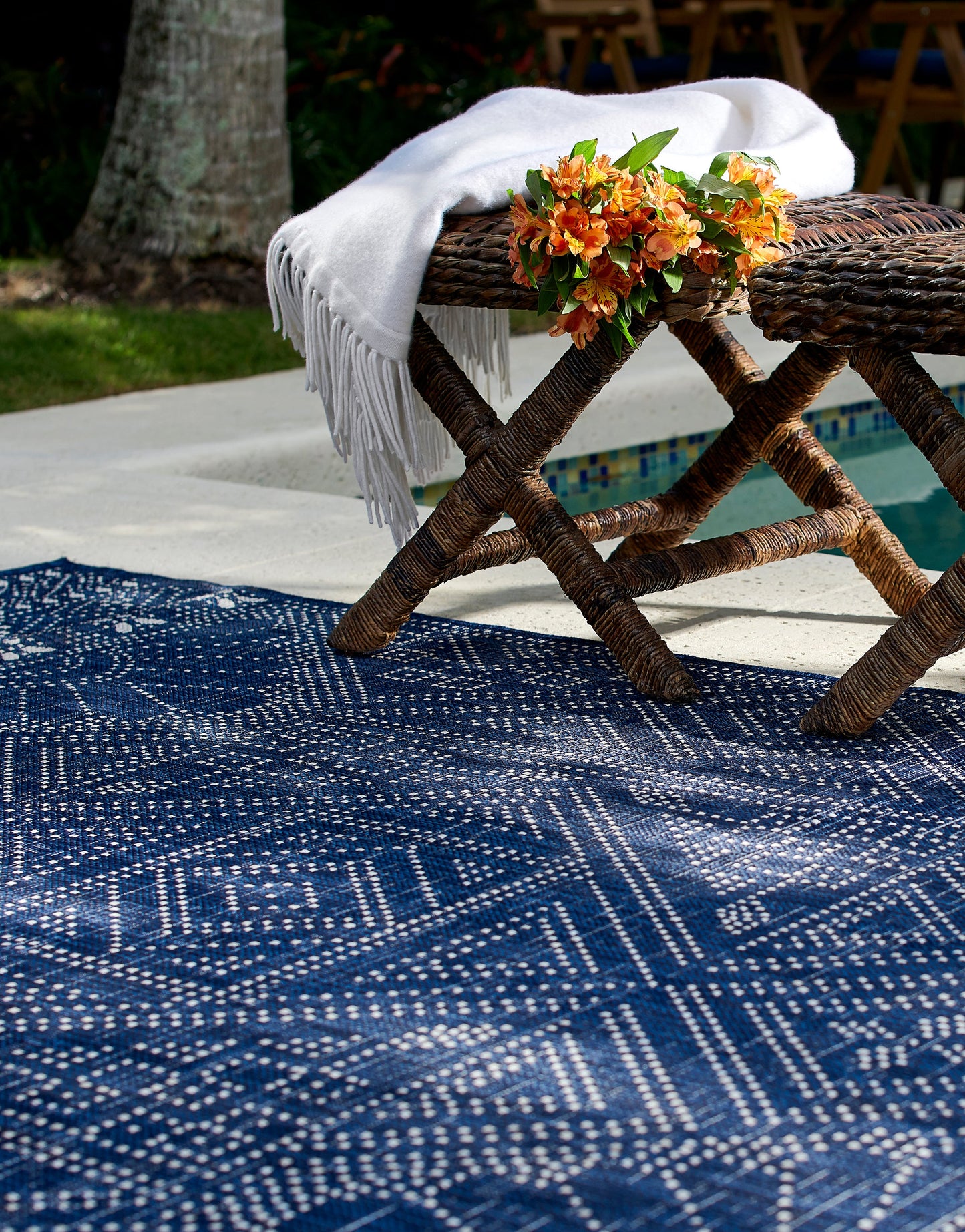 Outdoor patio scene: blue geometric rug, rustic cross-leg bench, white fringed throw, and orange bouquet by the pool.