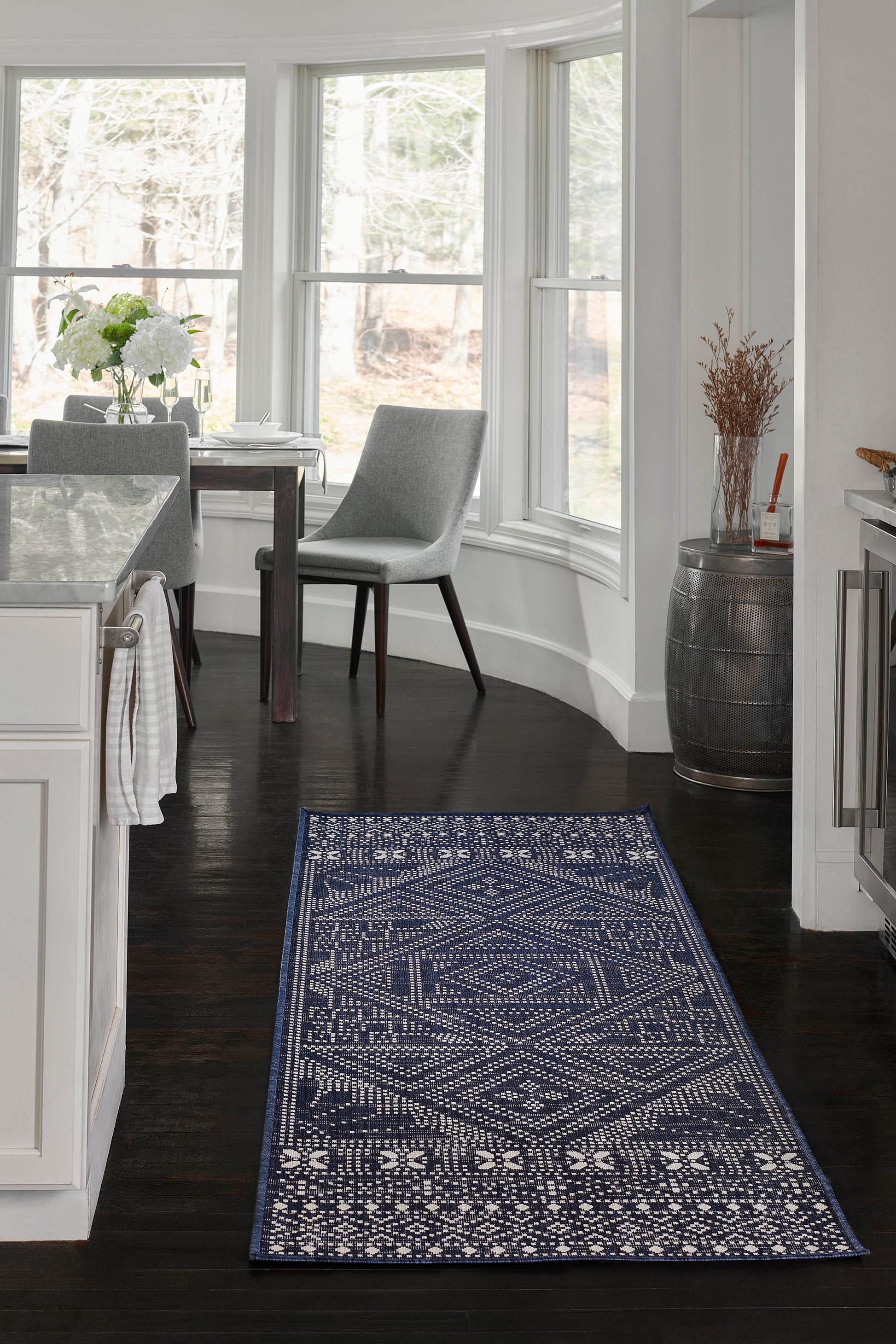 Navy blue geometric area rug on dark wood floor in a bright white kitchen-dining area with grey chairs.