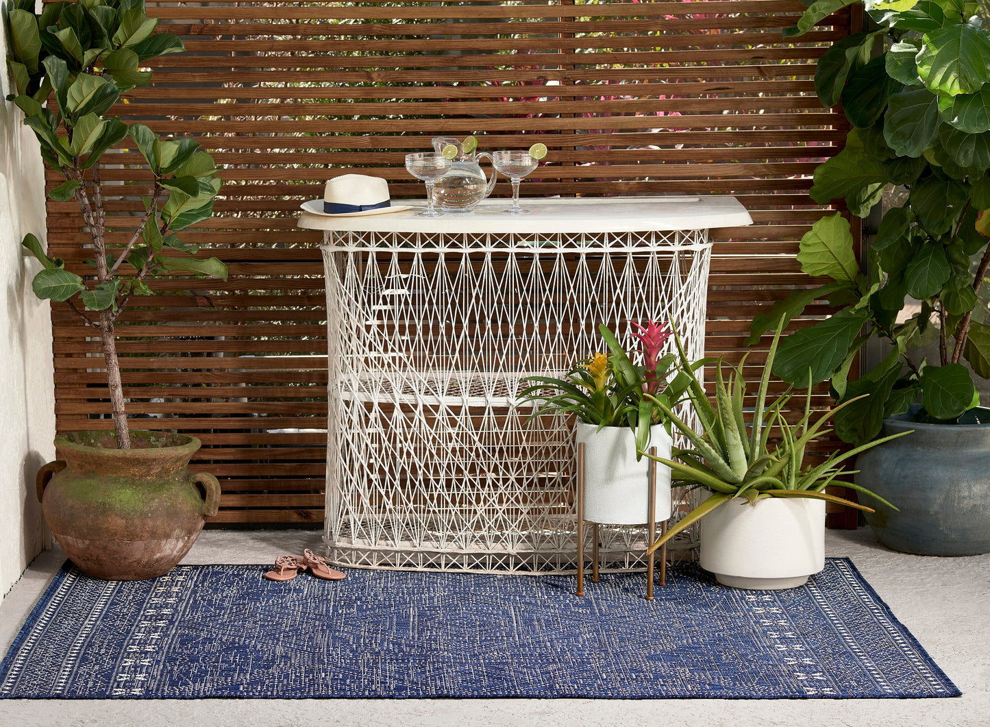 Blue outdoor rug with glassware & a hat on top, surrounded by potted plants.