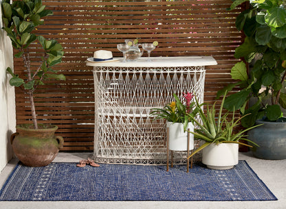 Navy outdoor rug under a white lattice-woven bar cabinet; potted plants, straw hat, margarita glasses on patio.
