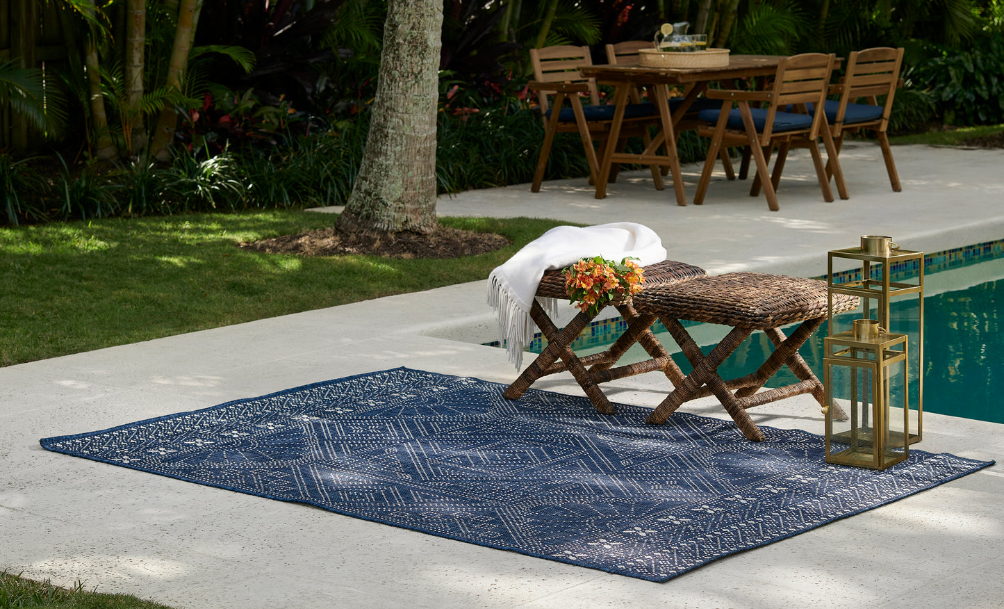 Blue geometric outdoor rug on a poolside patio with a wooden folding bench, woven table, white towel, and gold lanterns.