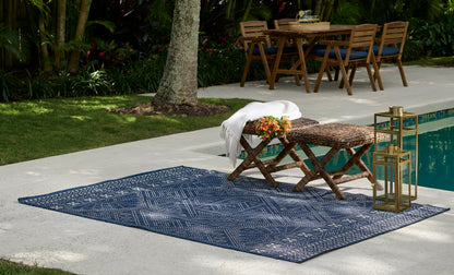 Blue geometric outdoor rug on a poolside patio with a wooden folding bench, woven table, white towel, and gold lanterns.