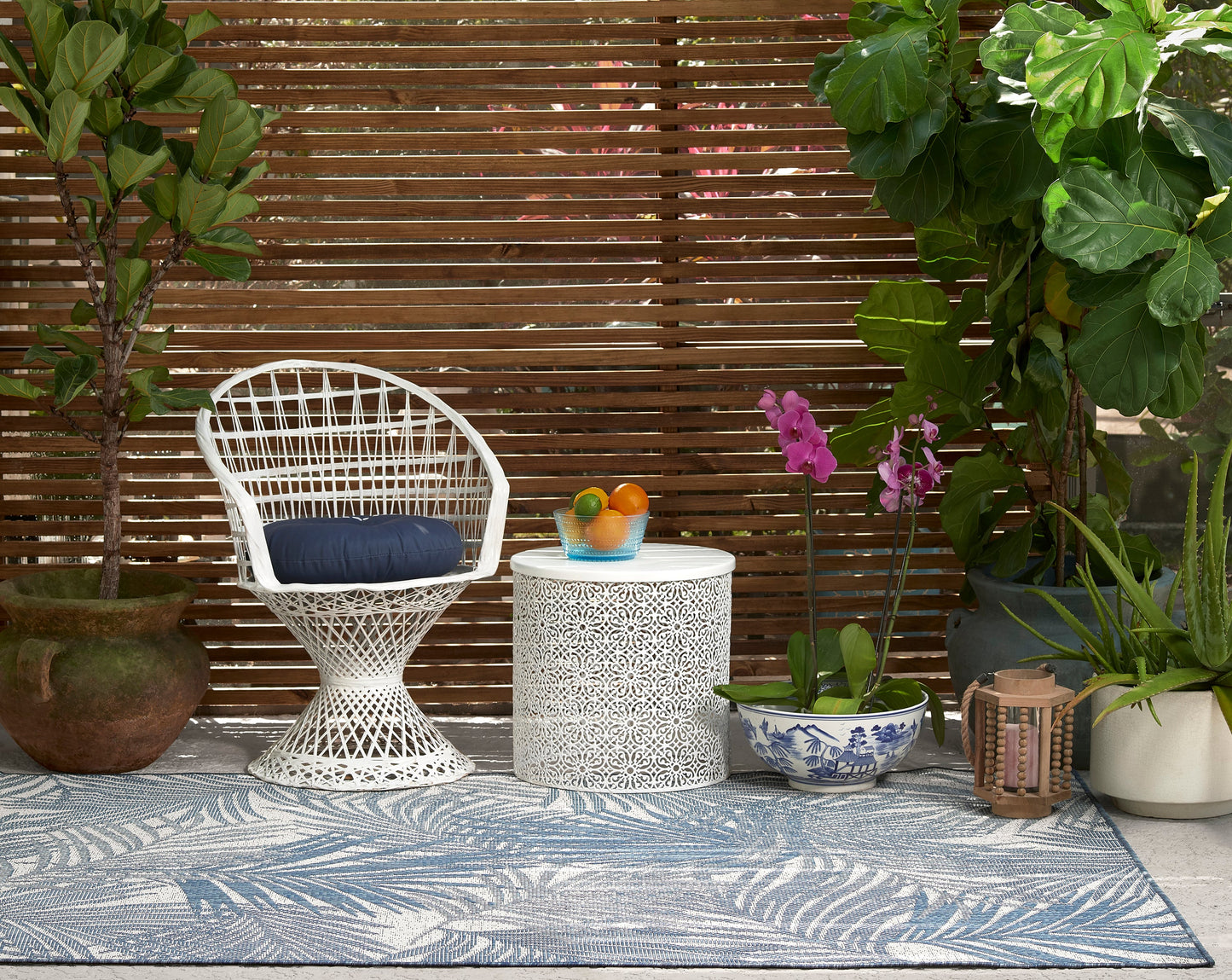 White wicker chair with navy cushion beside a white side table, fruit bowl, and potted plants on a blue patterned outdoor rug.