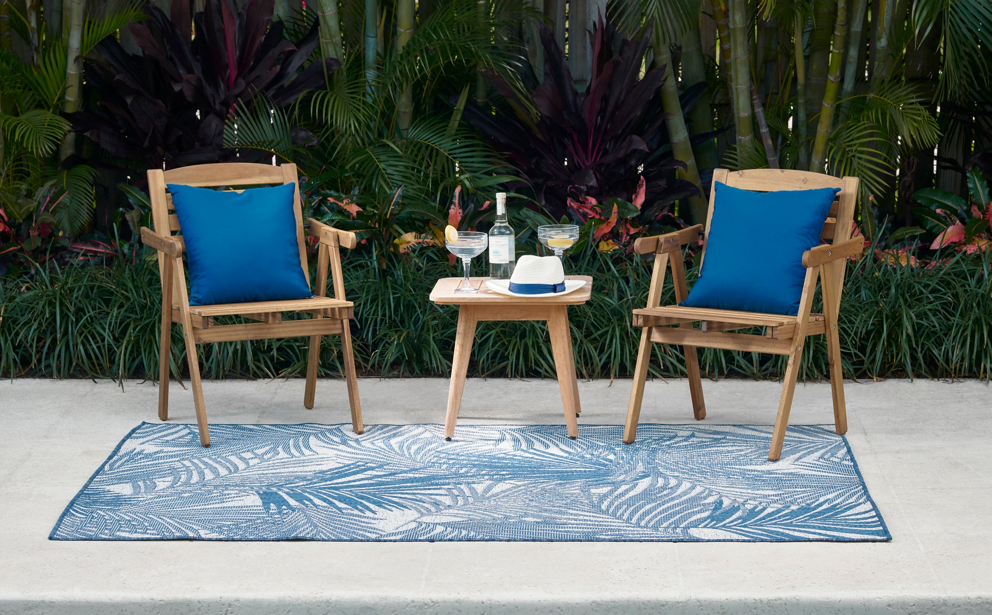 Two wooden patio chairs with blue cushions and a small round table between them on a palm-leaf patterned blue outdoor rug.