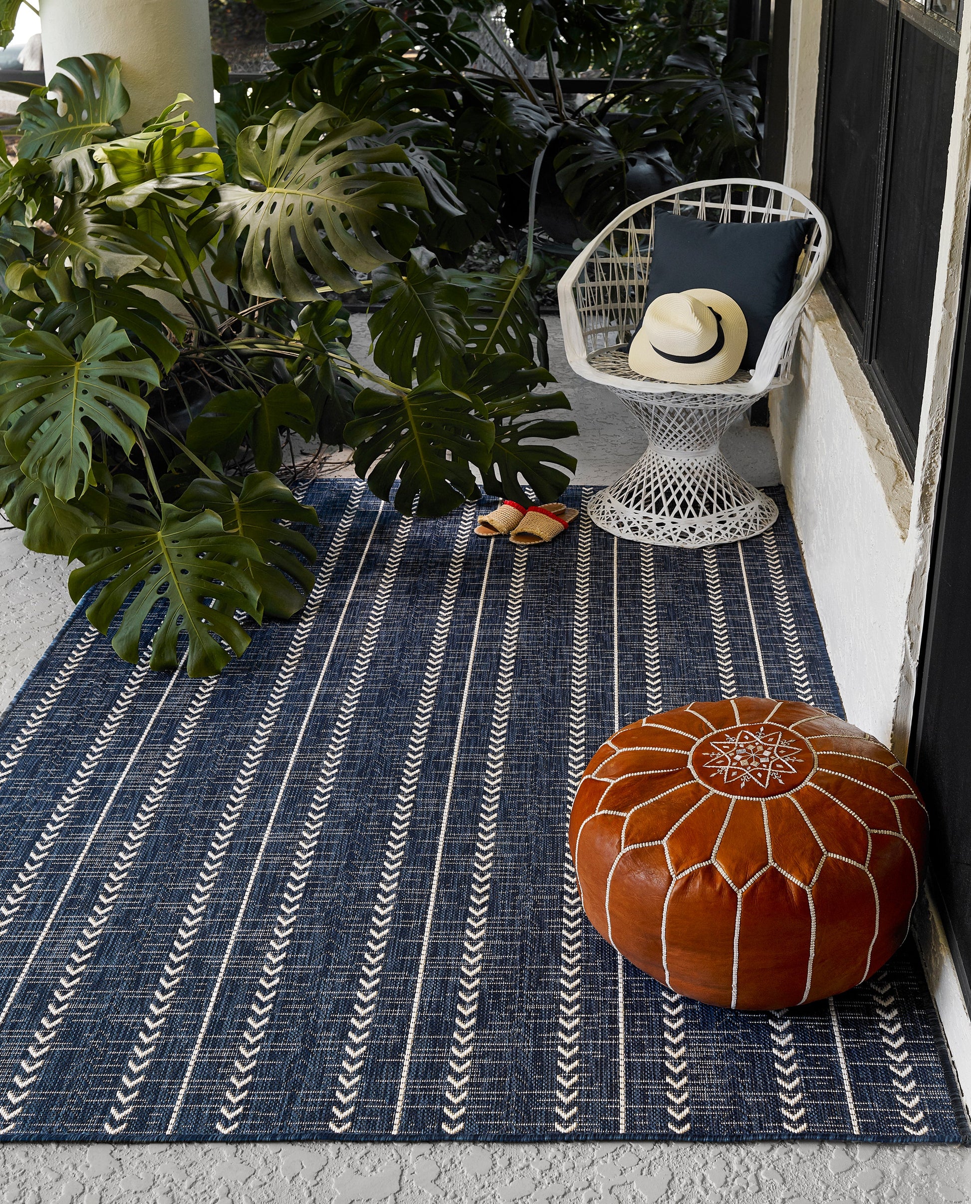 Navy striped outdoor rug with white lines, white wicker chair with dark cushion and hat, orange pouf and sandals nearby.