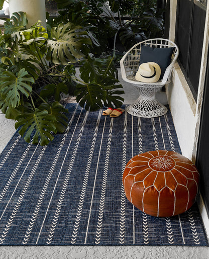 Outdoor rug scene: blue striped rug, white wicker chair, navy cushion, straw hat, orange stitched leather pouf.