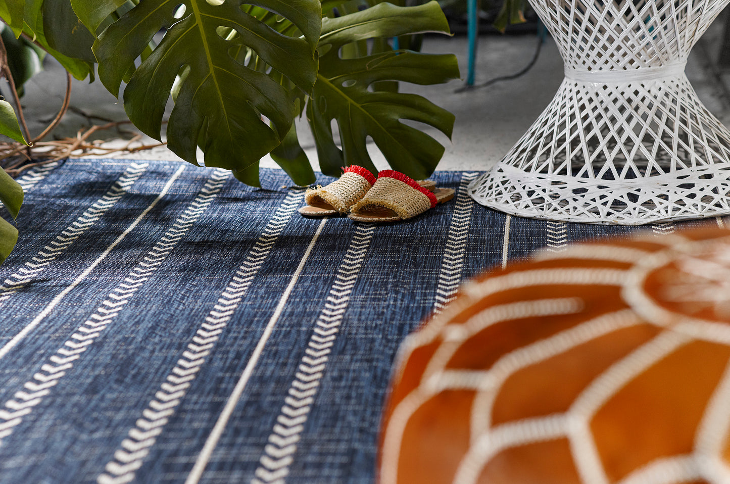 Navy striped outdoor rug with a monstera leaf, white wicker lamp, tiny straw sandals, and an orange pumpkin decoration.