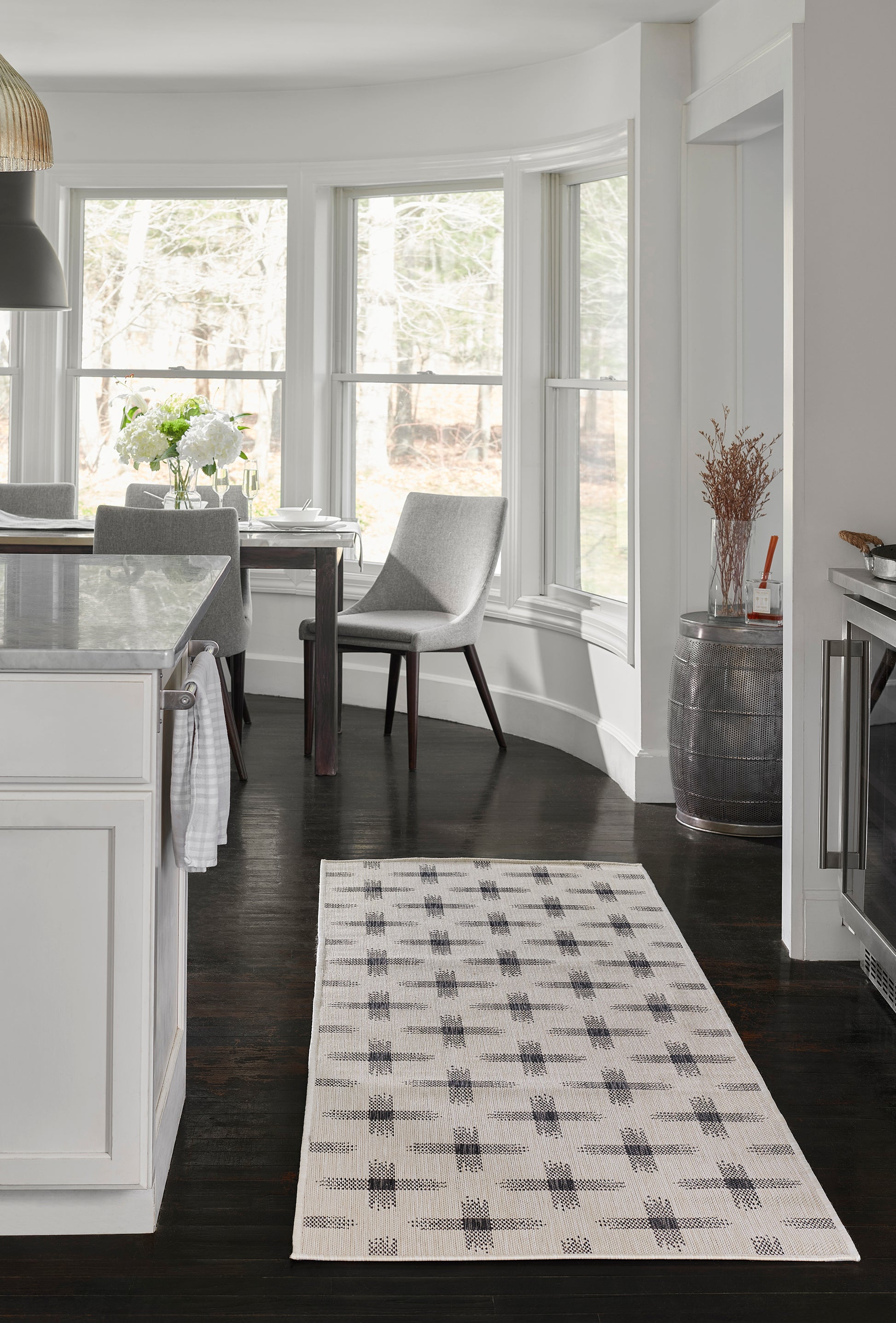 Modern kitchen with white cabinets, dark wood floors, marble island, windows, gray dining chairs, and a beige plaid runner.