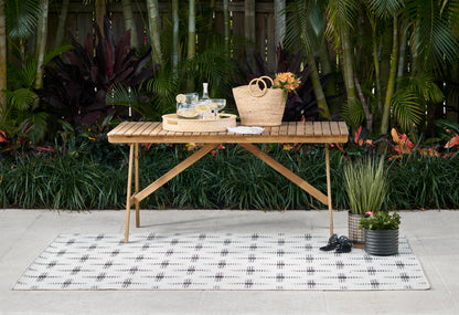 Outdoor rug with a grid pattern; a wood folding table sits on it, with a straw tote, glassware, bottle, and tropical plants nearby.