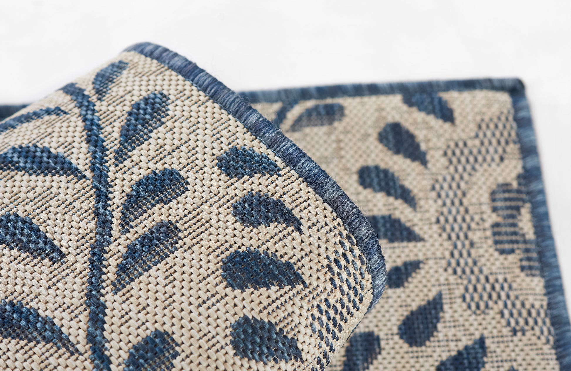 Beige woven rug with blue botanical leaf pattern; rolled corner shows textured weave and blue border.