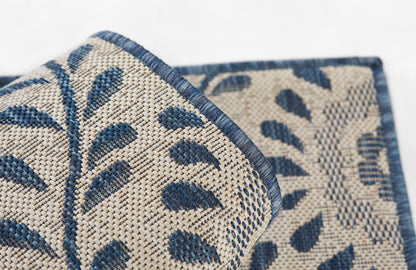 Beige woven rug with blue botanical leaf pattern; rolled corner shows textured weave and blue border.