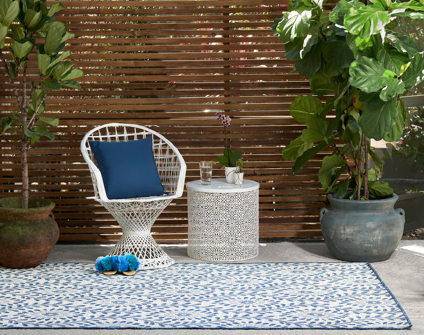 White wicker chair with blue cushion, white lattice side table, blue patterned outdoor rug, potted tropical plants.