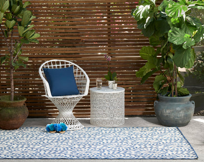 White wicker chair with blue cushion, white lattice side table, blue patterned outdoor rug, potted tropical plants.