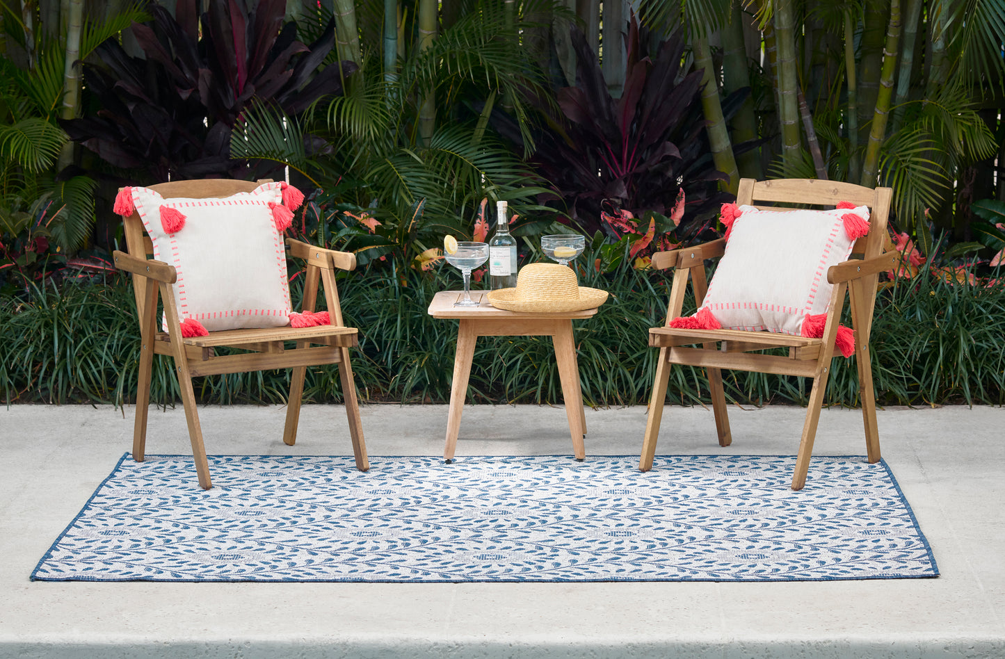 Outdoor patio set: two wood chairs with white cushions and red tassels, small table with drinks and a straw hat on a blue rug