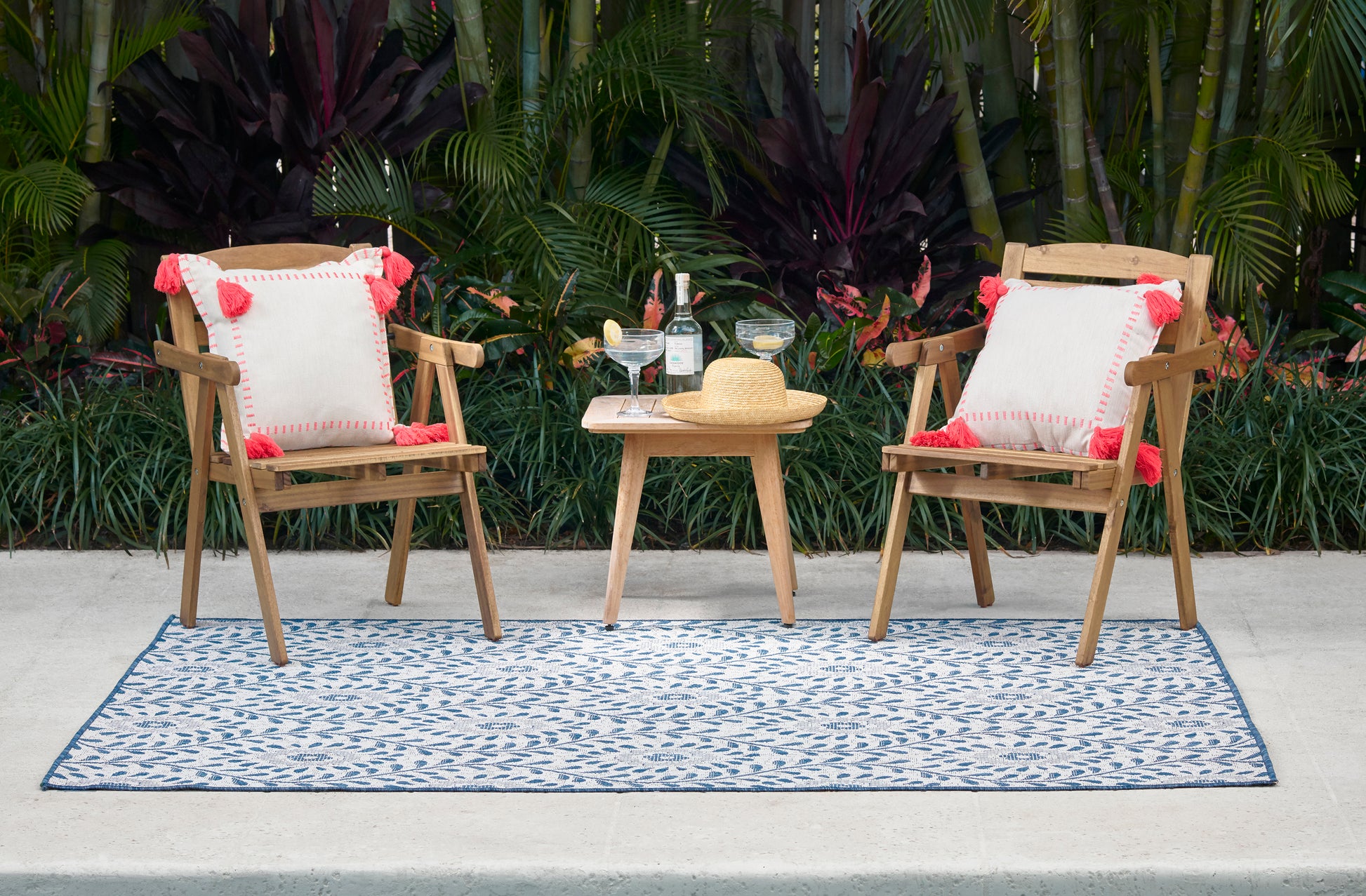 Outdoor patio set: two wood chairs with white cushions and red tassels, small table with drinks and a straw hat on a blue rug