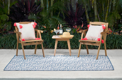 Outdoor patio set: two wood chairs with white cushions and red tassels, small table with drinks and a straw hat on a blue rug